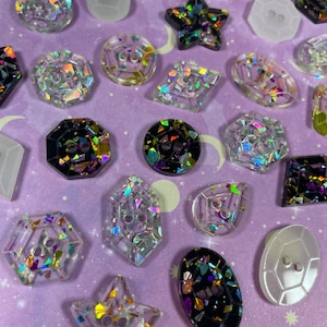 May include: Assortment of colourful resin buttons with various shapes and glitter accents. The buttons are in a variety of colours, including black, white, and clear. Some of the buttons are star-shaped, while others are round, square, or hexagonal.