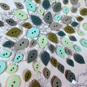 May include: A collection of leaf-shaped buttons in various shades of green, ranging from pale mint and sage to deep olive and forest green. Each button features two central holes for attachment, arranged in a vertical line on the surface.