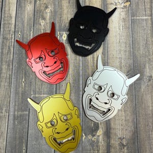 May include: Four colorful Oni mask bottle openers in red, black, yellow, and white. Each mask features horns and a wide, toothy grin. The bottle openers are arranged on a wooden surface.