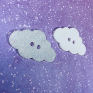 May include: Two white cloud-shaped buttons with glitter on one. The buttons have two holes each.