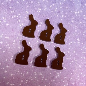 May include: Six brown wooden bunny buttons with two holes each. The buttons are arranged in two rows of three. The buttons are on a purple background with white stars.