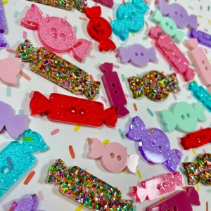 May include: Assortment of colorful candy-shaped buttons with glitter accents. The buttons are made of resin and come in various colors, including pink, red, purple, blue, and green.