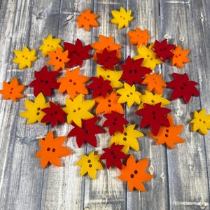 May include: A collection of colorful fall leaf-shaped buttons in shades of red, orange, and yellow. The buttons are arranged in a loose pile on a wooden surface.