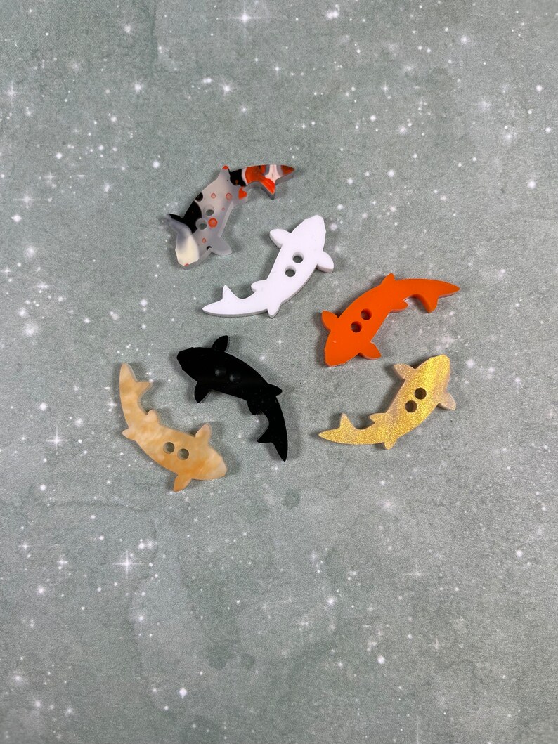 May include: Six colourful fish-shaped buttons with two holes each. The buttons are made of plastic and have a glossy finish. The colours include black, white, orange, and brown.