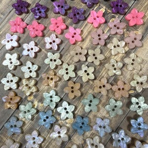 Peut inclure: Collection de boutons en forme de fleurs de différentes couleurs, dont rose, violet, blanc et bleu. Les boutons ont deux ou trois trous pour la couture et sont disposés sur une surface en bois.