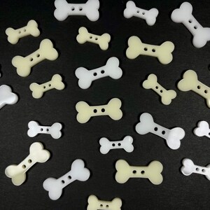 May include: A collection of white and off-white bone-shaped buttons. The buttons are arranged in a heart shape on a black background.