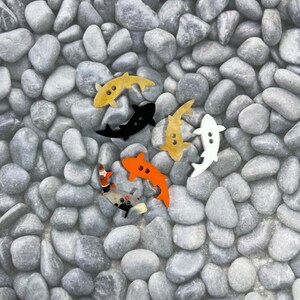May include: Five colourful wooden fish buttons on a background of grey stones. The fish are in various colours, including black, orange, yellow, and white.