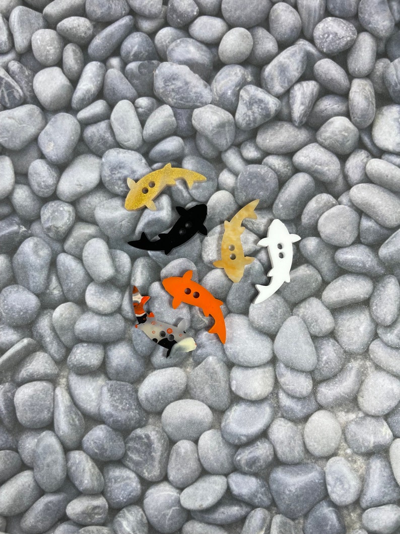 May include: Five colourful wooden fish buttons on a background of grey stones. The fish are in various colours, including black, orange, yellow, and white.