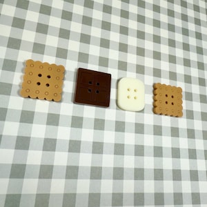 May include: Four decorative buttons in the shape of cookies and a chocolate bar. The buttons are in shades of brown and white, with a square, rectangular, and scalloped edge design. The buttons are arranged on a gray and white checkered background.