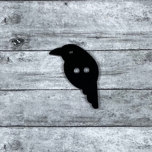 May include: Black wooden crow silhouette with two small holes. The crow is on a gray wooden background.