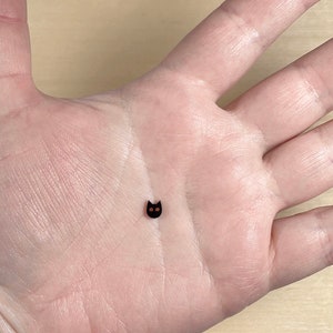 May include: A small black cat charm with orange eyes. The charm is on a person's hand.