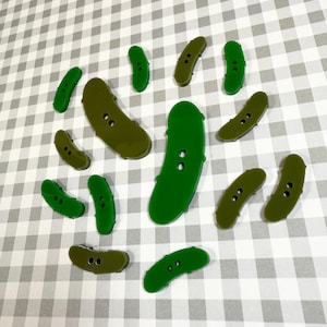 May include: A collection of pickle-shaped buttons in shades of green and olive. The buttons vary in size and have two or three holes for sewing. They are scattered on a gray and white checkered background.