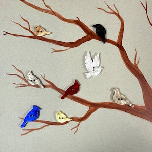 May include: A collection of bird-shaped buttons in various colors, including blue, red, black, white, and yellow, are arranged on a painted tree branch against a gray background. The buttons are made of a shiny material and have two or three holes for sewing.