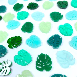 May include: Assortment of green, blue, and white monstera leaf shaped acrylic buttons. The buttons are various shades of green, blue, and white.