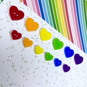 May include: A set of ten rainbow heart-shaped buttons with two holes each. The buttons are arranged in a rainbow order from red to purple, with the largest button at the top and the smallest button at the bottom.