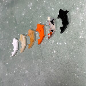 May include: A set of six colourful fish-shaped buttons. The buttons are made of plastic and have a glossy finish. The colours of the buttons are white, beige, orange, black, and a mottled black and white.