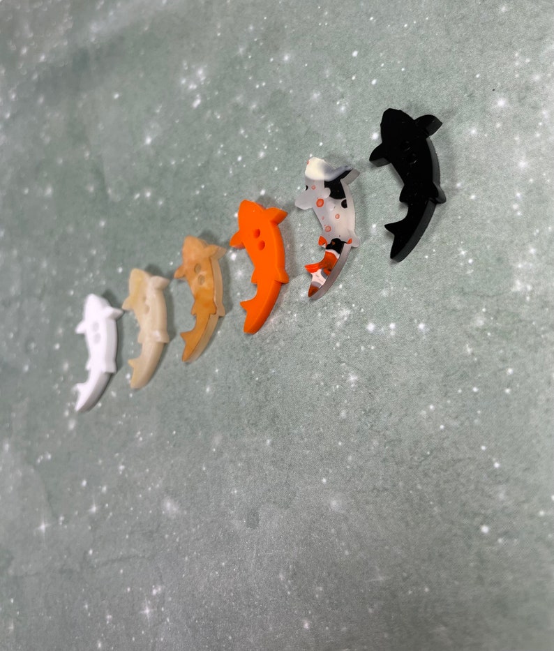May include: A set of six colourful fish-shaped buttons. The buttons are made of plastic and have a glossy finish. The colours of the buttons are white, beige, orange, black, and a mottled black and white.