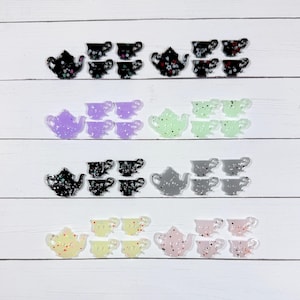 May include: A collection of small, flat teapot and teacup shaped charms arranged in four horizontal rows. The charms are made of translucent and opaque acrylic in various colours including black, purple, green, grey, yellow, and pink, all featuring embedded glitter and confetti-like accents.