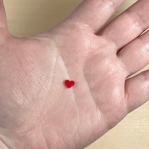 May include: A small, red heart-shaped bead resting on a person's palm.