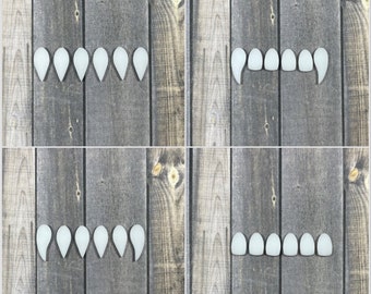 May include: Four sets of white plastic vampire teeth on a wood background. The teeth are arranged in a row, with the fangs on the outside and the other teeth in the middle.
