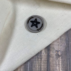 May include: A gray wooden button with a black star design and a brown leaf border.