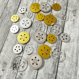 May include: A collection of circular buttons in various sizes and finishes, including solid gold, matte white, and transparent acrylic. Each button features a central five-pointed star design formed by thread holes, with some buttons having four or five holes.