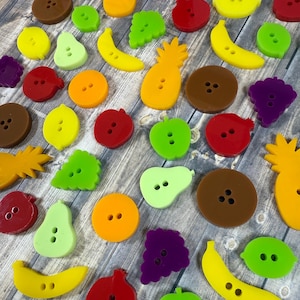 May include: A collection of colorful fruit-shaped buttons, including pineapples, bananas, pears, grapes, lemons, and apples. The buttons are made of plastic and have a smooth, glossy finish.