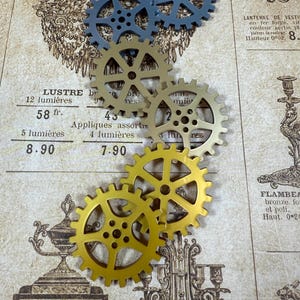 May include: An assortment of decorative gears in shades of gold and blue are arranged on a vintage-style patterned background. The gears vary in size and are made of metal. The background features French text and illustrations.
