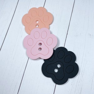 May include: Three paw print shaped buttons in pink, peach, and black. The buttons are made of a soft, flexible material and have two holes for sewing.