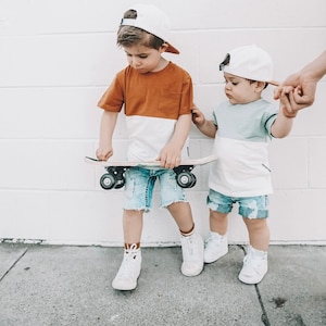 May include: Two young boys wearing white baseball caps and denim shorts. One boy is holding a small wooden skateboard. The boys are standing in front of a white brick wall.