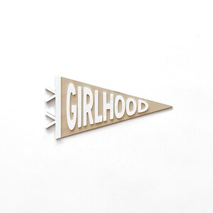 May include: A pennant-shaped sign with the word "GIRLHOOD" in white, three-dimensional letters. The pennant has a light brown background and white trim. The sign is set against a white background.
