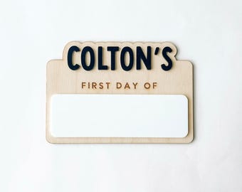May include: A light-colored wooden sign with a scalloped top edge featuring the black text COLTON'S and the engraved text FIRST DAY OF above a large, blank, rectangular white space designed for writing custom information.