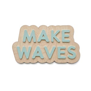 May include: Wooden sign with the words "MAKE WAVES" in light blue letters. The sign has a light brown wooden base with a wavy edge. The letters are raised and stand out against the background.