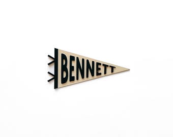 May include: A wooden pennant in a triangular shape with the name "BENNETT" in black letters. The pennant is light brown with black accents on the left side. The background is plain white.