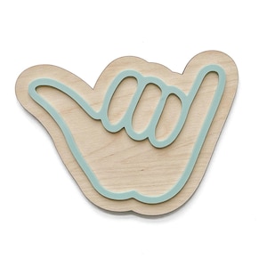 May include: Wooden wall art in the shape of a shaka hand gesture. The art piece features a light wood base with a light blue outline and interior details. The design is layered for a 3D effect, perfect for a coastal or surf-themed decor.
