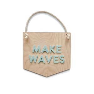 May include: A wooden sign with a rope hanger, painted in a light wood tone. The sign has the words "MAKE WAVES" in light blue letters.