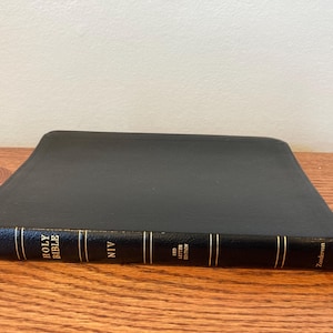May include: A black Bible with gold lettering and accents. The spine reads "Holy Bible NIV Red Letter Edition" and the publisher is Zondervan. The book is lying on a wooden surface.