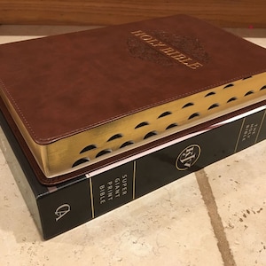 May include: A brown leather-bound Holy Bible with gold-gilded edges and a decorative design on the cover. The Bible is stacked on top of a black book with the text "The Holy Bible" and "Super Giant Print Bible" printed on the spine.