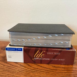 Könnte beinhalten: Eine in schwarzes Leder gebundene Life Application Studienbibel mit Großdruck, die auf ihrer originalen kastanienbraunen und weißen Pappschachtel liegt. Das Buch hat silberfarbene Seitenränder mit Griffregister, und der Titel steht auf der Schachtel.