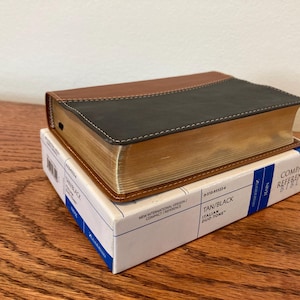 May include: A compact reference Bible in a tan and black leather cover, resting on its original box. The Bible's pages have a gold-colored edge. The box is white with blue accents and text that reads "Compact Reference Bible".