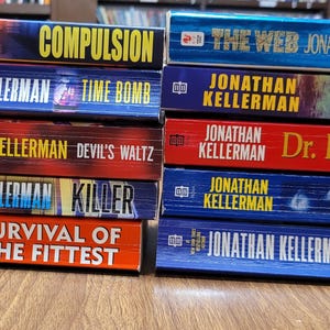 May include: A stack of Jonathan Kellerman books with titles like "Compulsion," "Time Bomb," and "The Web." The books have colorful covers, with titles in bold, contrasting fonts. The books are stacked on a wooden surface.