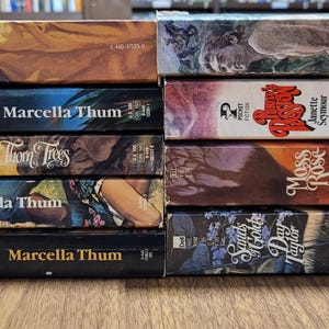 Könnte beinhalten: Ein Stapel alter Taschenbücher mit farbenfrohen Einbänden. Zu den Titeln gehören "A Rebel's Love", "Mistress of Paradise", "Thorn Trees", "Marguerite" und "Wild Laurel". Die Bücher sind auf einer Holzoberfläche gestapelt.