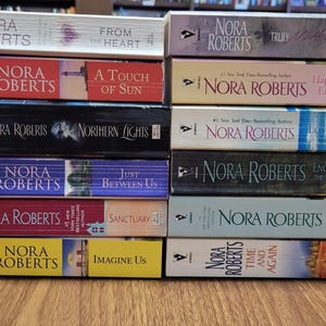 May include: A collection of Nora Roberts novels, featuring titles such as "Northern Lights," "Happy Endings," and "Time and Again." The books, with their colourful spines, are stacked on a wooden surface.