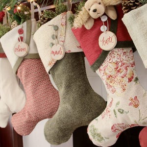 Puede incluir: Una fila de cinco calcetines navideños con texturas y patrones variados, que incluyen verde oliva sólido, estampados geométricos rojos y diseños florales. Cada calcetín está adornado con una etiqueta de madera redonda con los nombres oliver, madi o kate.