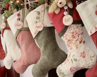 May include: A row of five Christmas stockings featuring varied textures and patterns, including solid olive green, red geometric prints, and floral designs. Each stocking is adorned with a round wooden name tag labeled oliver, madi, or kate.