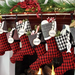 May include: A collection of Christmas stockings in red and black buffalo plaid, each personalized with a name tag. The stockings are hung above a fireplace, decorated with greenery and festive embellishments, creating a warm holiday scene.