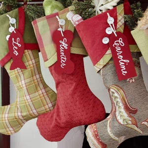 May include: Three Christmas stockings with personalized red tags. The first is green plaid with a fish-shaped tag labeled Leo. The second is red with a bone-shaped tag labeled Hunter. The third is a floral pattern with a rectangular tag labeled Caroline.