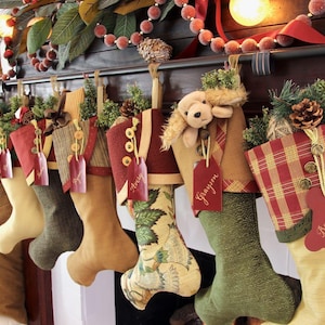 May include: A row of seven fabric Christmas stockings hanging from a dark wooden mantel. Each stocking features unique patterns, textures, and colors including red, green, tan, and plaid. Decorative tags with names like Avery, Grayson, and Bear are attached.