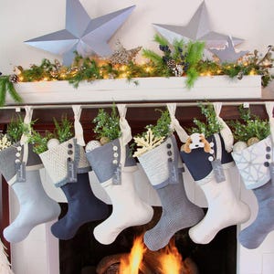 May include: Six fabric Christmas stockings hanging in a row from a metal rod. Each stocking features a unique combination of gray, navy blue, and white fabrics with textured patterns, decorative buttons, and small personalized name tags attached.