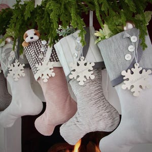 Puede incluir: Una fila de calcetines navideños en varios colores y patrones, colgados de una guirnalda verde. Cada calcetín tiene una etiqueta con forma de copo de nieve con un nombre. Un calcetín tiene un pequeño perro de peluche.
